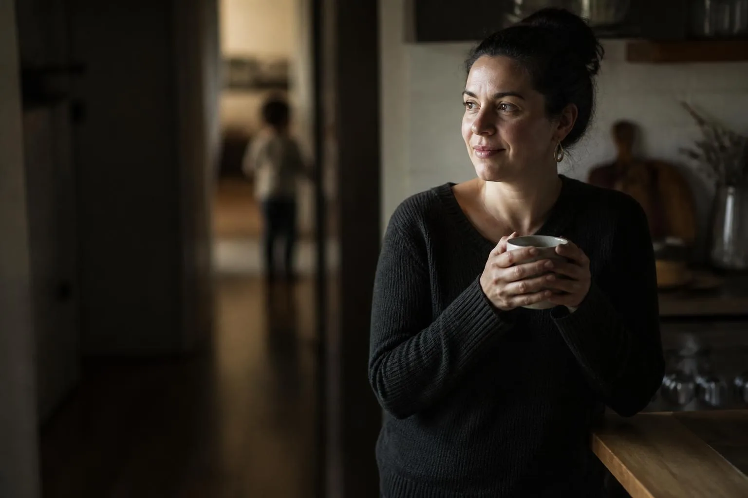 A parent holding a mug at a kitchen counter, reassured, a child out of focus behind.
