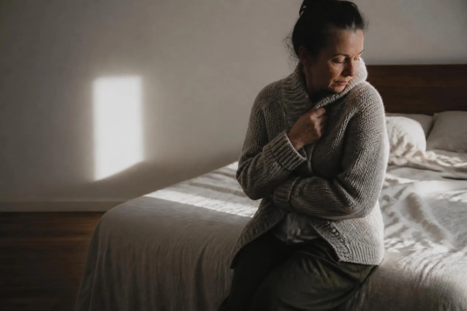 A woman on the edge of a bed pulling a wool cardigan around her in morning light.