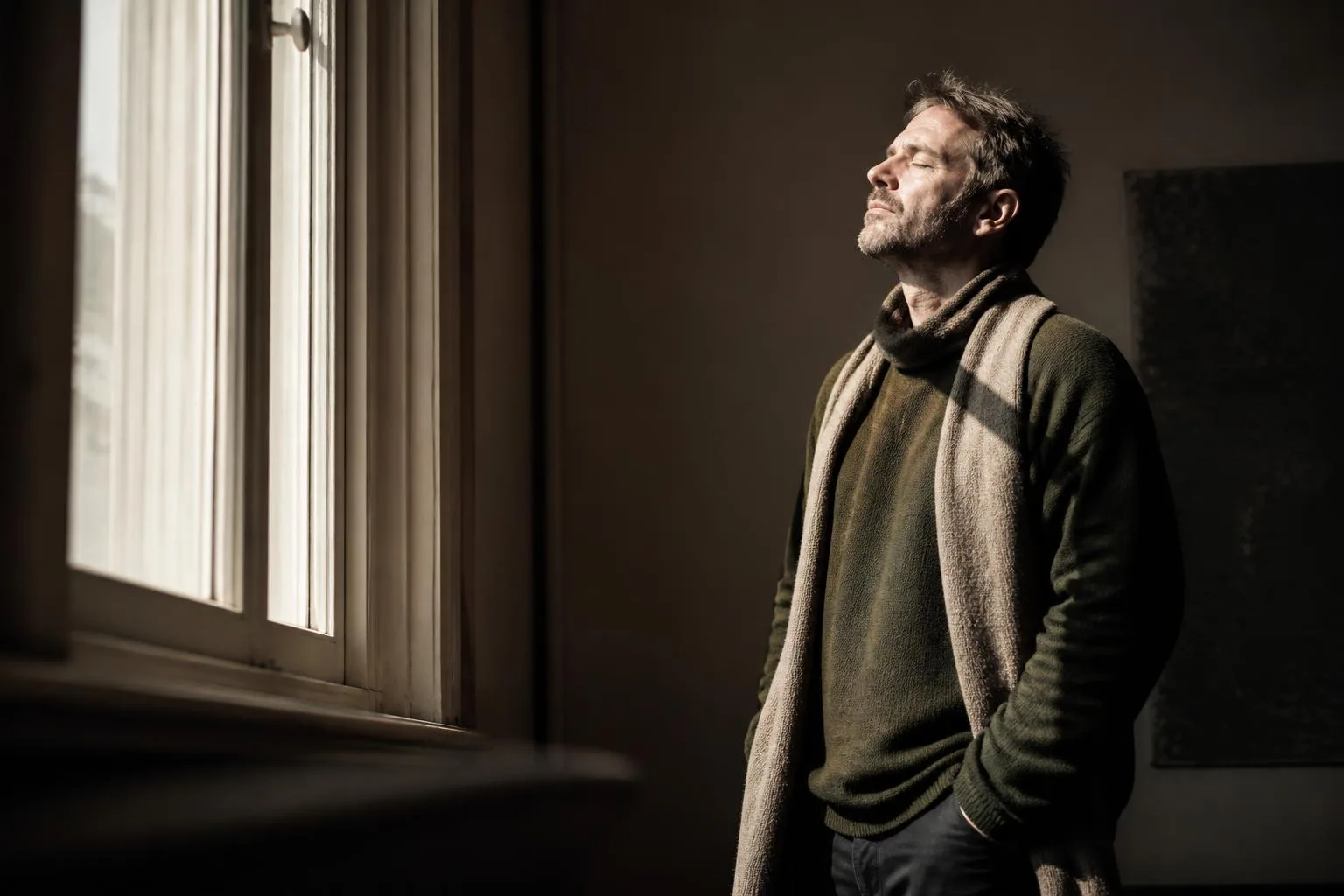 A man at a bright winter window, face turned fully into the daylight, eyes closed.