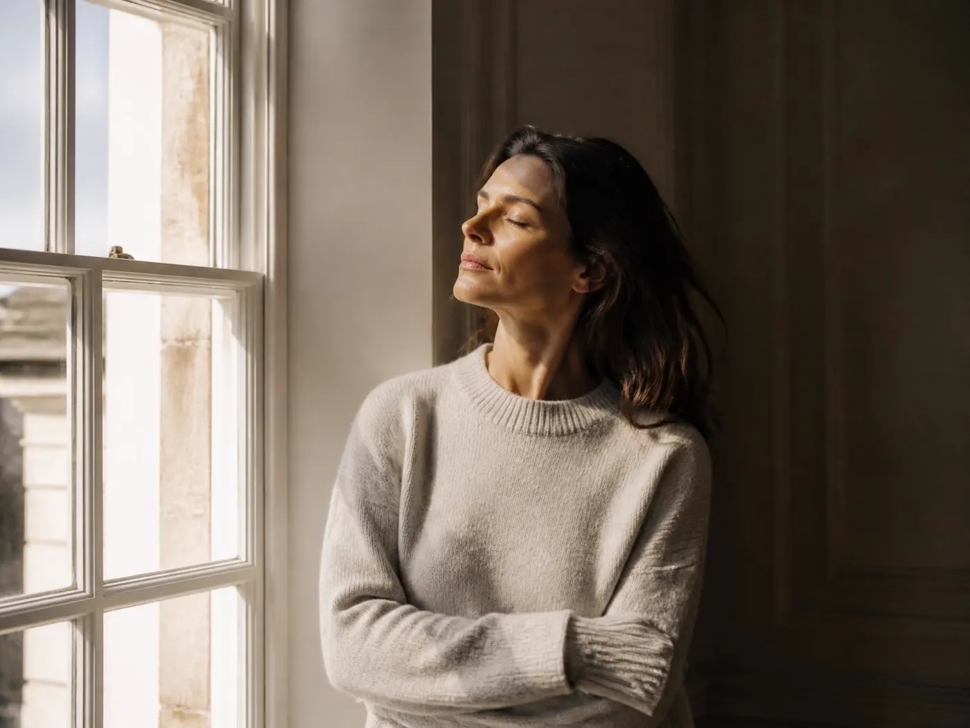 A woman standing at a tall sash window, face lifted into late-afternoon light.
