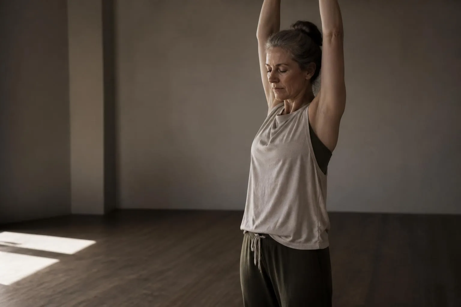 A woman in her fifties mid-stretch in a bare bright room, capable and strong.